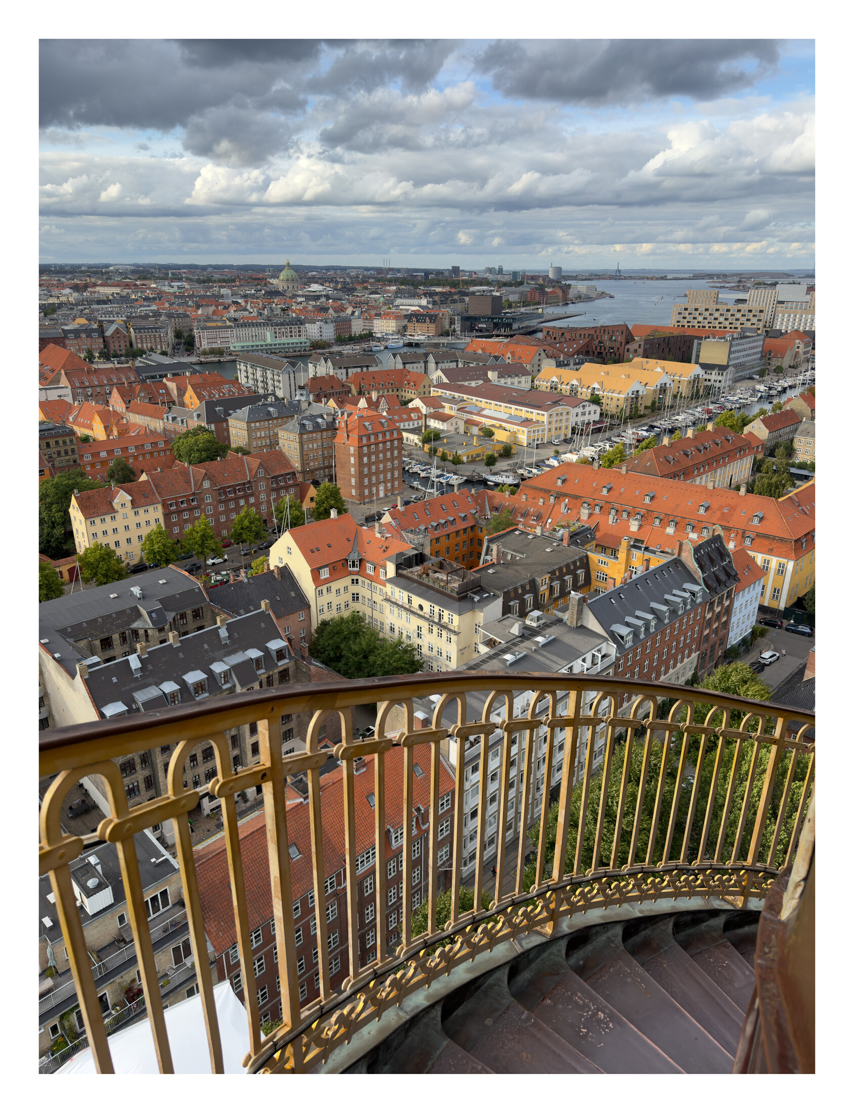 Copenhagen rooftops and harbor beyond the gold railing and exterior stairs of Vor Frelsers Kirke